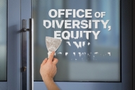 A person is scraping text that says “Office of Diversity, Equity and Inclusion” from a door.