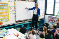 A young female teacher in a brightly decorated elementary classroom writes on a whiteboard, as the young students, sitting on the floor, pay close attention.