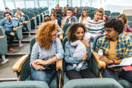 A young mother holds her baby in a lecture hall, with other students sitting around her.