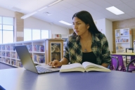 A student in a library works on her laptop with a book open next to her