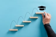 Male hand arranges a wooden block ladder with academic mortar board symbol at the top of the last step