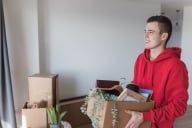 A young man in a red hoodie carries moving boxes into an empty apartment
