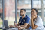 A small group of medical students are seen sitting at desks in a classroom during a lesson. They are each wearing scrubs and paying close attention.