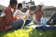 Three Black students sit on a lawn smiling. Two high five each other.