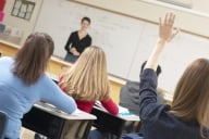 View of back of students in class with one student raising her hand before a professor at a white board