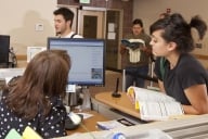 A student leans over a desk to look at a computer screen alongside a faculty member 