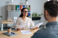 Woman at office desk in front of bookcase smiles as she talks to young man, whose head you see from the back