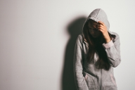 A college student stands in a poorly lit room with hood on her head and hand over her face, looking embarrassed.