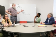 Mark Hoyert stands at a whiteboard with a group of five faculty members who are focused, considering his words
