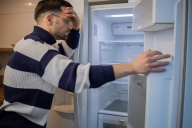 A young man holds a fridge door open and leans against the fridge, his head in his hand, as he surveys its contents: the fridge is empty. 