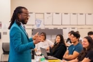 A dark-skinned man with shoulder-length hair wearing a blue jacket shares a story with students in his class, some of whom are smiling.