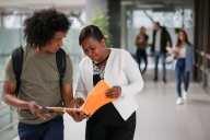 Happy teacher talking to an African American college student in the hall at the school and pointing at his notebook