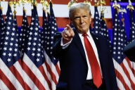 Donald Trump, in a suit and red tie, stands in front of a row of American flags and points his finger.