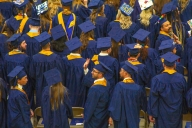 A group of students in blue and yellow graduation regalia face away from the camera. One student turns to the left to wave to someone out of frame.
