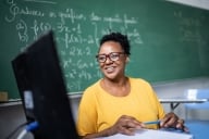 Middle aged woman with dark skin and dark hair and glasses sits in front of a computer screen with a chalkboard with math problems behind her.
