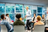 Young man in casual clothes stands in the middle of a seated circle of peers and a facilitator. 