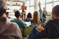 A female student, seated in a college classroom and pictured from the back, raises her hand to respond to a prompt from the professor at the front of the class.