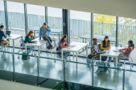 University students are seen scattered at various desks in an open study area, as they work on assignments between classes. Some students are working together while others are seen working independently in this aerial view.