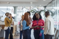 A diverse group of young professionals stand chatting during a networking event; some are carrying water bottles and some are wearing lanyards with name tags.