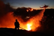 A firefighter battles a fire while a helicopter drops water on the blaze