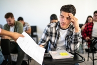 A young man with his fingers on his forehead prepares to take a graded assignment back from a professor.
