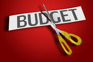 A pair of scissors cuts through a piece of paper with the word "BUDGET," against a red background.