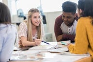Four college students sit around a table, one of them with a pencil in her hand as if she is explaining something.