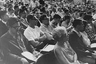 In this historical black-and-white photo from a 1965 anti-war teach-in, a dense crowd of people in an auditorium sit in an audience, seemingly listening to a speaker with intense interest and focus.
