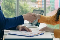 Two people shaking hands in front of a laptop. The laptop is open and has a picture of a person on it.