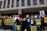 A person in a black shirt holds a megaphone in front of the Education Department building, surrounded by yellow signs and other people