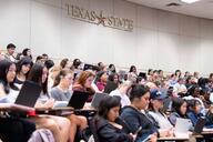 A group of students sit in a large lecture hall, facing the front of the room. Behind them on a large white wall is the Texas State logo.