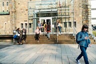 Medium group of multiethnic students going in and out of a college building entrance. Some are sitting and standing on a small staircase on a sunny day.
