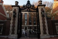 A photo of a student walking through campus gates at Harvard University, a stately brick campus building in the background.
