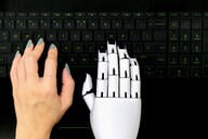 Human hand and a robot hand both hover over a computer keyboard. 