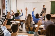 A classroom teacher stands at the head of a roomful of students who are raising their hands.