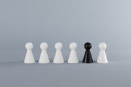 A row of six pawns (chess pieces) against a gray background. Five of the pawns are white and one pawn, placed second to the right, is black.