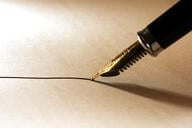 A close-up photo of a fountain pen with black ink being wielded to draw a thin line on paper.