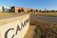 A wall with word “campus” written on it can be seen with an academic building and open space in the distance.