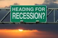 A green and white road sign, as if above a highway, says "Heading for Recession?" against the backdrop of a sunset in a cloudy sky.