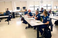 Students in scrubs and masks sit at desks listening to an instructor.