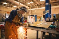 A young female student practices her welding skills in a technical classroom. She wears a face mask and sparks from the welding can be seen around her. 