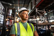 A Black woman in a hard hat and neon vest stands in a warehouse.