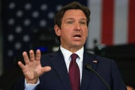 A photo of Florida Governor Ron DeSantis, a white man with brown hair, wearing a dark suit with a maroon tie and gesturing with his right hand.