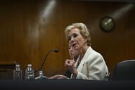 Linda McMahon in a white blazer sits at a table in a hearing room in the Capitol