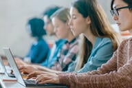 A line of college students of different races and ethnicities works on their laptops in a classroom.