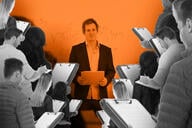 A professor with a large group of people holding clipboards around them as they teach.