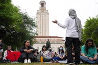 A woman wearing a kaffiyeh as a head scarf addresses a crowd of students seated in the grass in front of a building at the University of Texas at Austin.
