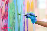 A photo of an artist's gloved hand holding a paintbrush and painting a brightly colored picture.