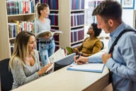 A smiling female librarian engages with a student checking out a book, while two other students can be seen talking in the background.