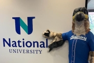 A person wearing a falcon mascot costume poses next to a sign reading "National University."